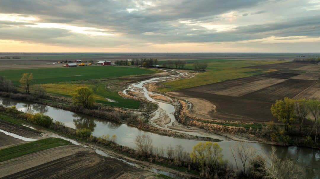 A photograph of a sprawling rural farmland viewed from directly overhead, showcasing the stark impact of agricultural runoff. A prominent channel of muddy, brownish water snakes from freshly plowed fields directly into a slow-moving river, visibly discoloring the water near the point of entry. Scattered across the landscape are patches of vibrant green crops, bare soil, and a few distant red barns and yellow tractors, all set against a backdrop of a diffused, overcast sky with subtle hints of sunlight breaking through. The scene conveys a sense of scale and highlights the environmental consequences of intensive farming practices.